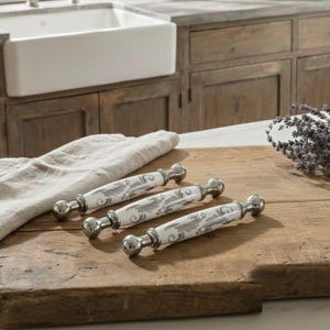 Può includere: Quattro pomelli decorativi per mobili con manici in ceramica bianca con un motivo floreale grigio ed estremità in metallo argentato lucido. I pomelli sono disposti su un tagliere di legno, suggerendo un tema di cucina o di arredamento per la casa.