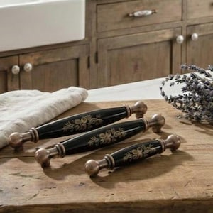 May include: Three black cabinet pulls with bronze-colored ends and floral designs. The pulls are arranged on a wooden surface, with a white sink and wooden cabinets in the background. A bundle of lavender is also present.