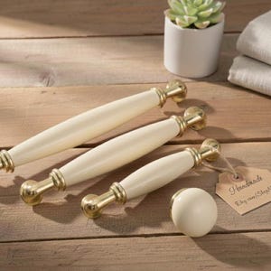 Peut inclure: Tirants de meubles crème avec des accents dorés, disposés sur une surface en bois. L'ensemble comprend trois tirants et un bouton assorti. Une petite étiquette indique "Handmade".
