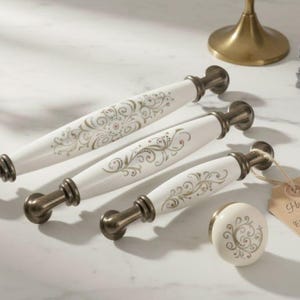 Puede incluir: Un juego de cuatro tiradores de cerámica blanca y dorada y un pomo a juego. Los tiradores tienen un diseño floral y herrajes metálicos de color bronce. Se adjunta una etiqueta que dice "Handmade".