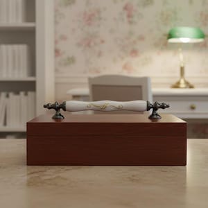May include: A rectangular, dark brown wooden box with a white and gold handle. The handle is centered on the top of the box and is flanked by ornate, dark metal hardware. The box is sitting on a light-colored surface.