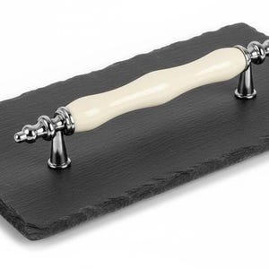 May include: A rectangular black slate serving board with a cream-colored handle. The handle is made of a glossy, curved material with silver-colored metal accents. The board has a textured surface and is suitable for serving appetizers.