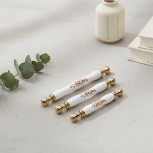 Könnte beinhalten: Drei weiße Keramik-Schrankgriffe mit goldfarbenen Enden und floralen Designs in Orange, Blau und Grün. Die Griffe sind auf einer hellgrauen Oberfläche angeordnet, mit einer kleinen Vase und Büchern im Hintergrund.