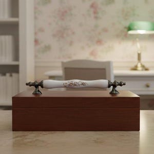 Puede incluir: Una caja rectangular de madera marrón oscuro con un asa decorativa blanca y dorada. El asa tiene detalles ornamentados y está montada sobre herrajes de color bronce antiguo. La caja está sobre una superficie clara.