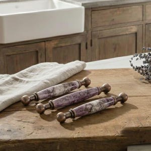 May include: Three cabinet pulls with a marble-effect purple and white design and bronze-coloured ends are displayed on a wooden surface. A white sink and wooden cabinets are in the background. A linen towel and lavender sprigs are also present.