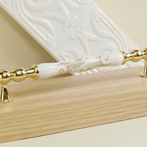 Può includere: Maniglia decorativa per mobili con centro in ceramica bianca con dettagli floreali dorati. La maniglia ha dettagli in metallo dorato ed è montata su una superficie in legno chiaro. Per mobili.