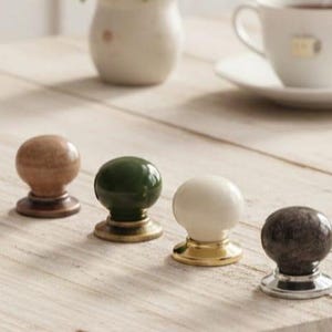 May include: Four decorative cabinet knobs in various colors and finishes. The knobs include a brown wooden knob, a green knob with a brass base, a white knob with a gold base, and a dark gray knob with a silver base.