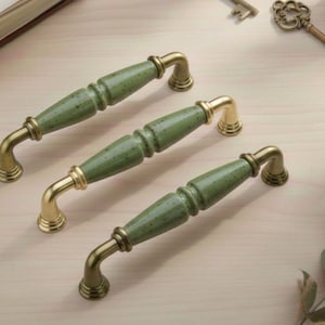 May include: Three cabinet pulls with a speckled green ceramic centre and gold-toned metal hardware. The pulls are arranged on a light-coloured wooden surface, showcasing their design and finish.