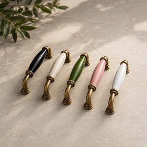May include: Five cabinet pulls with brass-colored hardware and ceramic handles in black, white, green, pink, and white. The pulls are arranged in a row, showcasing their different colors and designs. The handles are oval-shaped.