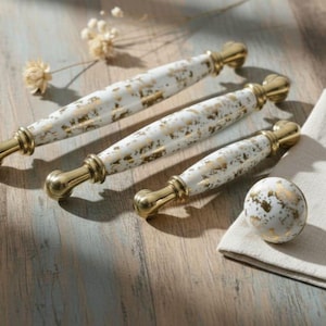 May include: A collection of cabinet hardware, including three white and gold speckled handles and a matching knob. The handles have a brass finish on the ends. The hardware is arranged on a light wood surface.