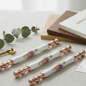 Może przedstawiać: Trzy białe i miedziane uchwyty do szafek z kwiatowymi wzorami. Uchwyty mają biały ceramiczny środek z kolorowym kwiatowym wzorem i miedzianymi metalowymi końcami. Uchwyty mają około 15 cm długości.