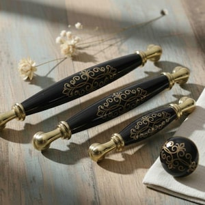 以下が含まれることがあります： 黒と金のキャビネットハードウェアのセット。セットには、3つのハンドルとノブが含まれています。ハンドルは、金色のアクセントと装飾的なデザインが施された黒いボディです。ノブは金色のデザインの黒色です。木製の表面にハードウェアがあります。