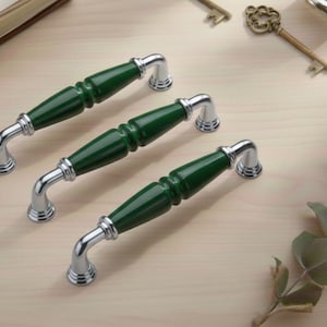 May include: Three cabinet pulls with a dark green, cylindrical center and chrome-colored curved handles. The pulls are arranged on a light-colored wooden surface, suggesting a home decor theme.