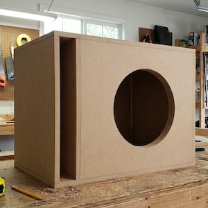 Puede incluir: Una caja de altavoz de madera sin terminar con una abertura circular y un puerto rectangular. La caja está hecha de madera de color marrón claro y está sobre un banco de trabajo de madera. Se ven herramientas y suministros en el fondo.