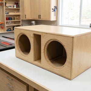 Puede incluir: Una caja de altavoz de madera sin terminar de color claro con dos grandes aberturas circulares y una abertura rectangular. La caja es rectangular y se encuentra sobre una superficie blanca en un taller. El taller tiene herramientas y armarios.