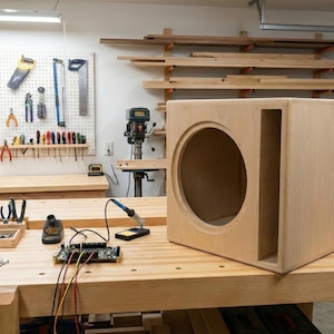Könnte beinhalten: Eine hellfarbene Holzlautsprecherbox mit einer großen kreisförmigen Öffnung und einem rechteckigen Anschluss. Die Box steht auf einer Werkbank in einer Werkstatt, umgeben von Werkzeugen und Geräten.