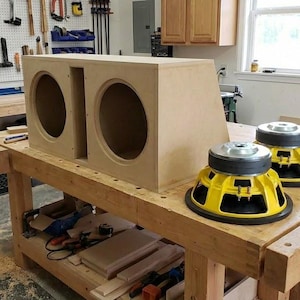 Könnte beinhalten: Ein hölzernes Subwoofer-Gehäuse mit zwei kreisförmigen Öffnungen steht auf einer Werkbank. Zwei gelb-schwarze Subwoofer befinden sich neben dem Gehäuse. Die Werkstatt hat Werkzeuge und Geräte an den Wänden und Werkbänken.
