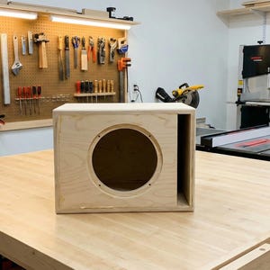 May include: A light-colored, unfinished wooden speaker box with a circular opening. The box is rectangular with an open side. It sits on a large wooden workbench in a workshop setting, with tools and equipment visible in the background.