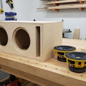 Op de afbeelding: Een houten luidsprekerbox met twee cirkelvormige uitsparingen en een rechthoekige poort, samen met twee zwarte en gele luidsprekers. De items staan op een houten werkbank in een werkplaats.