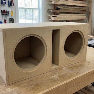 Puede incluir: Una caja de altavoz rectangular de madera sin terminar con dos grandes recortes circulares y un divisor central. La caja, hecha de madera de color claro, está sobre un banco de trabajo de madera en un taller. Diseñada para equipos de audio.