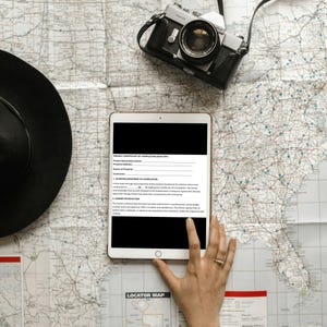 May include: A black vintage camera with a leather strap rests on a detailed map, next to a black hat and a tablet displaying a document. A hand with a gold ring is holding the tablet. The map has a locator map.