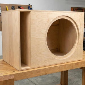 Puede incluir: Una caja de altavoz de madera clara sin terminar, con una gran abertura circular y un puerto rectangular. La caja está construida con paneles de madera y descansa sobre un banco de trabajo de madera. Se ven herramientas en el fondo.