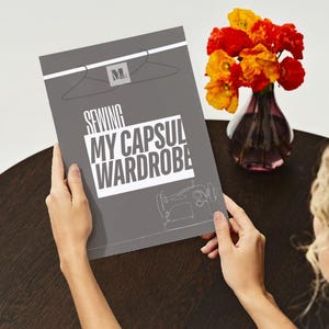 Könnte beinhalten: Eine Person hält ein graues Buch mit dem Titel "Sewing My Capsule Wardrobe". Das Cover zeigt einen Kleiderbügel, eine Nähmaschinenillustration und die Initialen "ML". Eine Vase mit orangefarbenen und roten Blumen steht in der Nähe auf einem dunkelbraunen Tisch.