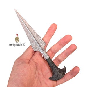 Può includere: Un pugnale argentato e nero con una lama e un'impugnatura ornate. La lama presenta un motivo dettagliato e l'impugnatura presenta un design decorativo. Il testo "erfolgsDRUCK" e un logo sono visibili sullo sfondo.