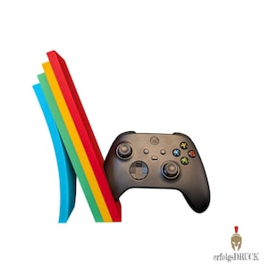 Könnte beinhalten: Ein schwarzer Xbox-Controller lehnt an einer farbenfrohen, gebogenen Struktur aus Blau, Grün, Gelb und Rot. Der Controller hat mehrere Tasten und Joysticks. Der Hintergrund ist weiß.