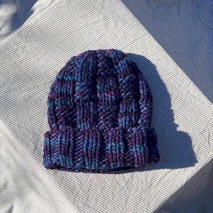 May include: A hand-knitted beanie hat in shades of purple, blue, and teal. The hat features a ribbed knit design and a folded brim. The hat is made of thick, chunky yarn.