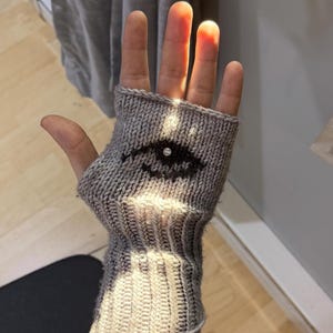 May include: Gray knitted fingerless glove with a black eye design. The glove is made of a textured knit fabric and features a ribbed cuff. The eye design is centered on the back of the hand.
