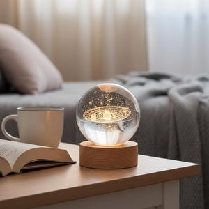 Puede incluir: Una esfera de vidrio transparente con un diseño iluminado del sistema solar, colocada sobre una base redonda de madera. La esfera está iluminada desde dentro, mostrando los planetas y las estrellas. La lámpara está sobre una mesita de noche de madera.
