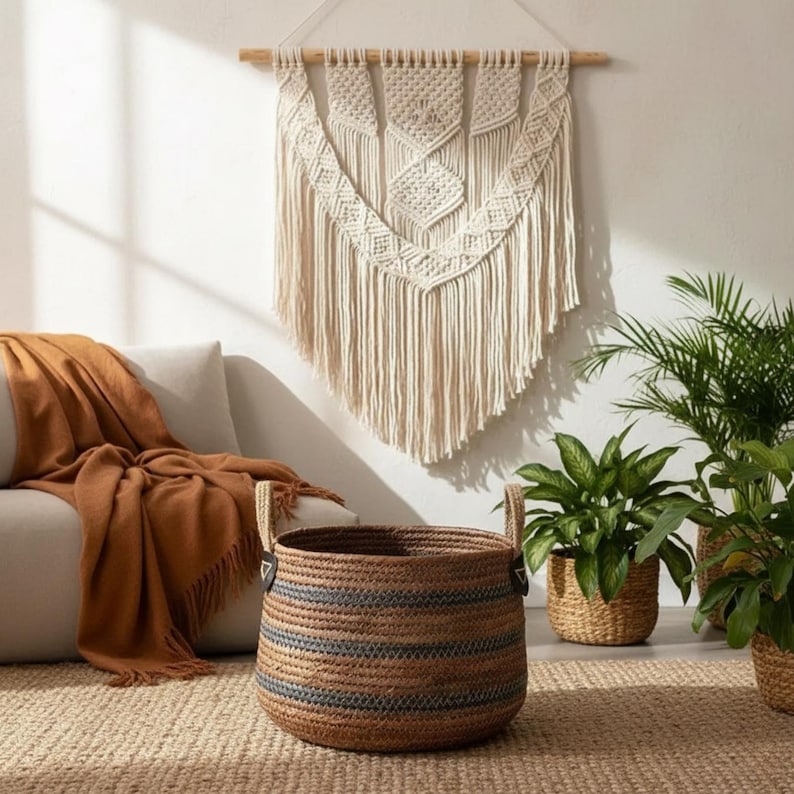 May include: A woven basket with dark blue stripes and handles sits on a jute rug. A beige macrame wall hanging with diamond patterns and fringe hangs above a sofa with a brown throw blanket. Potted plants are in the background.