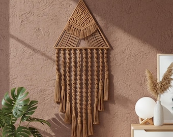 Puede incluir: Un colgante de pared de macramé marrón en forma triangular. La decoración de pared presenta intrincados detalles de nudos y flecos, añadiendo un toque bohemio. La pieza está suspendida contra una pared texturizada de color marrón rojizo, creando un ambiente cálido.