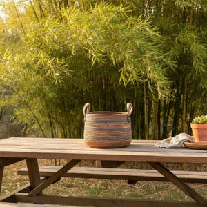 May include: A round woven basket with two handles, featuring alternating bands of brown and blue-gray. The basket sits on a wooden picnic table, with a bamboo background. A small terracotta pot with a plant is also on the table.