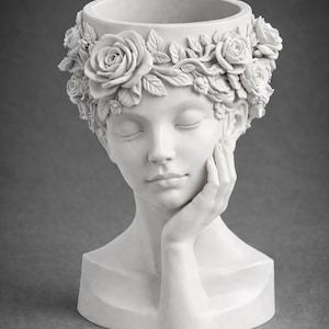 May include: A white ceramic planter in the form of a woman's head, featuring closed eyes and a hand touching her cheek. The head is decorated with a floral crown of roses and leaves. The planter has a wide opening at the top.