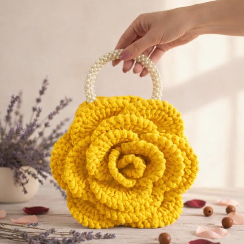 K&ouml;nnte beinhalten: Eine handgefertigte gelbe H&auml;keltasche in Rosenform. Die Tasche hat einen Griff aus wei&szlig;en Perlen. Die Tasche wird von einer Hand gehalten. Der Hintergrund umfasst Lavendel und Holzperlen.