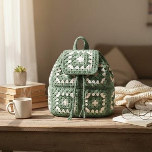 May include: A handmade green and white crochet backpack with a drawstring closure. The backpack features a granny square design and a top handle. It is sitting on a wooden table next to a stack of books and a coffee mug.