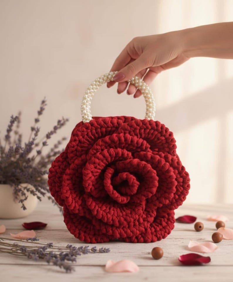 May include: A red crocheted handbag in the shape of a rose, featuring a handle adorned with pearls. The bag's petals are carefully crocheted, with a spiral centre. The handle is a curved arch of white pearls. A unique accessory.