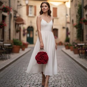 K&ouml;nnte beinhalten: Eine Frau in einem wei&szlig;en Kleid geht eine Kopfsteinpflasterstra&szlig;e entlang und h&auml;lt eine rote, rosenf&ouml;rmige Handtasche. Das Kleid hat einen V-Ausschnitt und Spaghettitr&auml;ger. Die Handtasche ist tiefrot und hat einen Perlenhenkel. Im Hintergrund sind Geb&auml;ude und Topfpflanzen zu sehen.
