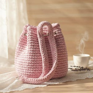 K&ouml;nnte beinhalten: Eine hellrosa geh&auml;kelte Bucket Bag mit Kordelzugverschluss und zwei langen Tr&auml;gern. Die Tasche ist aus dickem Garn gefertigt und hat ein strukturiertes Aussehen. Eine Tasse Kaffee und Lavendelzweige sind im Hintergrund.