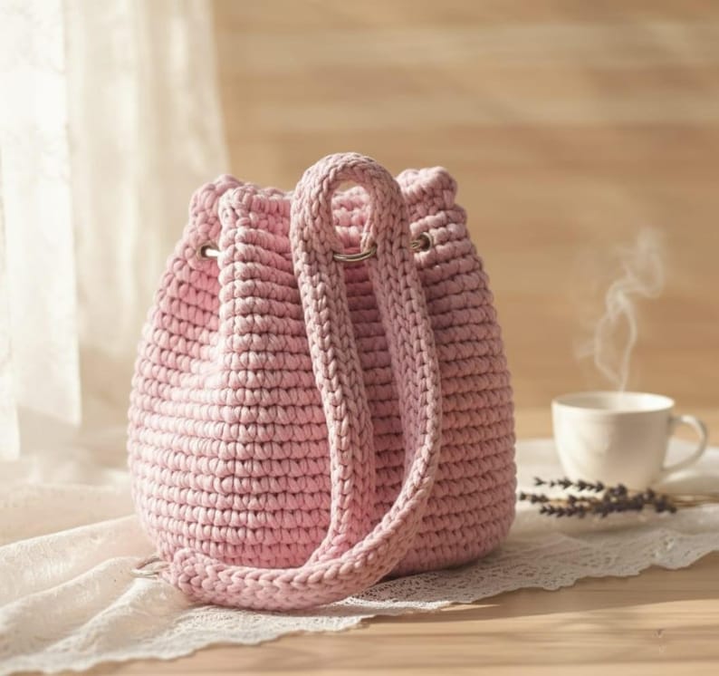 K&ouml;nnte beinhalten: Eine hellrosa geh&auml;kelte Bucket Bag mit Kordelzugverschluss und zwei langen Tr&auml;gern. Die Tasche ist aus dickem Garn gefertigt und hat ein strukturiertes Aussehen. Eine Tasse Kaffee und Lavendelzweige sind im Hintergrund.