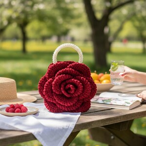 K&ouml;nnte beinhalten: Eine handgefertigte, tiefrote H&auml;keltasche in Rosenform mit einem wei&szlig;en Perlenhenkel. Die Tasche steht auf einem Picknicktisch aus Holz mit einem wei&szlig;en Tuch, Himbeeren und einem Strohhut. Der Hintergrund ist ein verschwommener gr&uuml;ner Park.