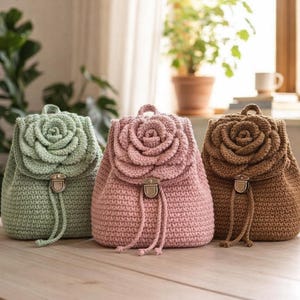 May include: Three crochet backpacks in green, pink, and brown, each with a large rose design on the flap. The bags have drawstring closures and metal hardware, displayed on a wooden surface.