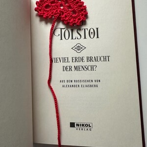 Könnte beinhalten: Ein rotes, gehäkeltes herzförmiges Lesezeichen in einem Buch. Das Buch ist auf einer Seite mit dem Namen des Autors, TOLSTOI, und dem Titel "VIEVIEL ERDE BRAUCHT DER MENSCH?" geöffnet. Der Faden des Lesezeichens erstreckt sich bis zu einem kleineren Herzen.
