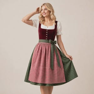 May include: A dirndl dress featuring a dark red velvet bodice with a row of silver buttons, a white blouse with short puffed sleeves, and a green skirt with a red and white gingham apron tied at the waist with a matching green fabric bow.