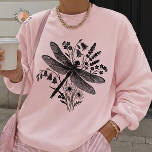 May include: A light pink sweatshirt featuring a black dragonfly and floral design. The graphic includes a dragonfly with detailed wings, surrounded by flowers and foliage. The sweatshirt has long sleeves and a crew neck.