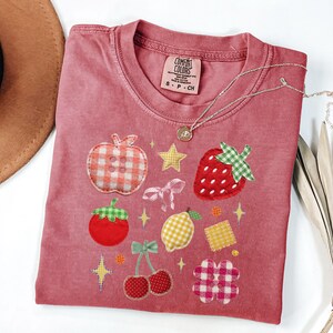 以下が含まれることがあります： 赤いComfort ColorsのTシャツに、果物と星のアップリケが施されています。アップリケには、リンゴ、イチゴ、サクランボ、レモン、花が含まれています。ペンダント付きのシルバーネックレスがシャツに付いています。