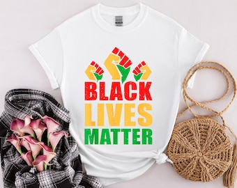 May include: White t-shirt with the text "BLACK LIVES MATTER" in red, yellow, and green. The design includes three raised fists in red, yellow, and green. A black and white plaid scarf and a woven bag are also in the image.