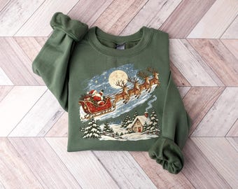 Könnte beinhalten: Ein olivgrünes Sweatshirt mit Rundhalsausschnitt und einem Grafikdruck, der den Weihnachtsmann in einem roten Schlitten zeigt, gezogen von Rentieren, die über eine verschneite Landschaft mit Haus und Tannen fliegen.